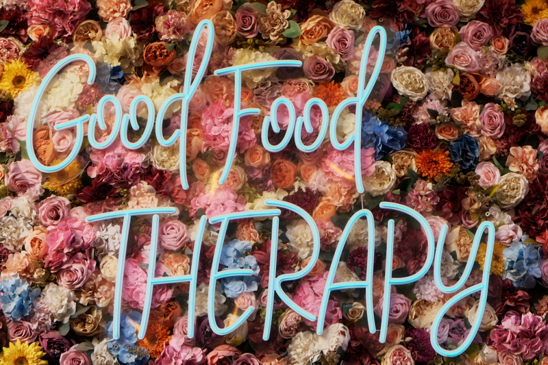 3 - Vibrant, aesthetic display, featuring a neon sign reading Good Food THERAPY mounted on a dense wall of mixed roses and flowers